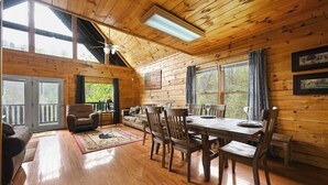 Interior de una cabaña de madera con comedor, sofá y ventanas amplias.