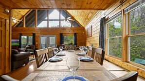 Una cabaña de madera con una mesa de comedor dispuesta para seis, un televisor y ventanas amplias con vistas al bosque.