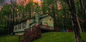 Une maison à deux étages avec un escalier en bois menant à une terrasse, entourée d’arbres et illuminée de guirlandes lumineuses.