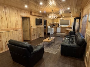 A log cabin interior with a living area featuring two black leather sofas, a wooden dining table, and a kitchen with a stove and microwave.