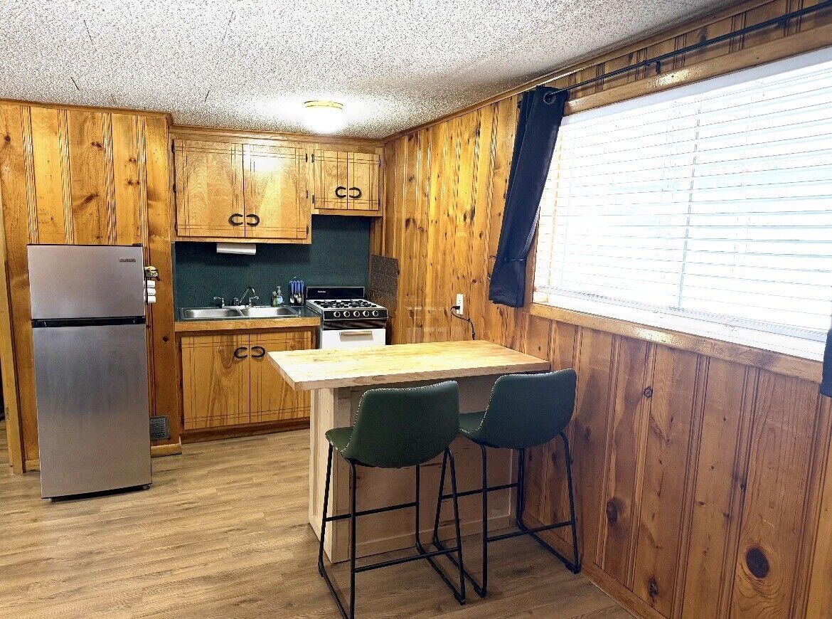 A compact kitchen with wooden cabinets, a refrigerator, a stove, and a bar area with two stools.