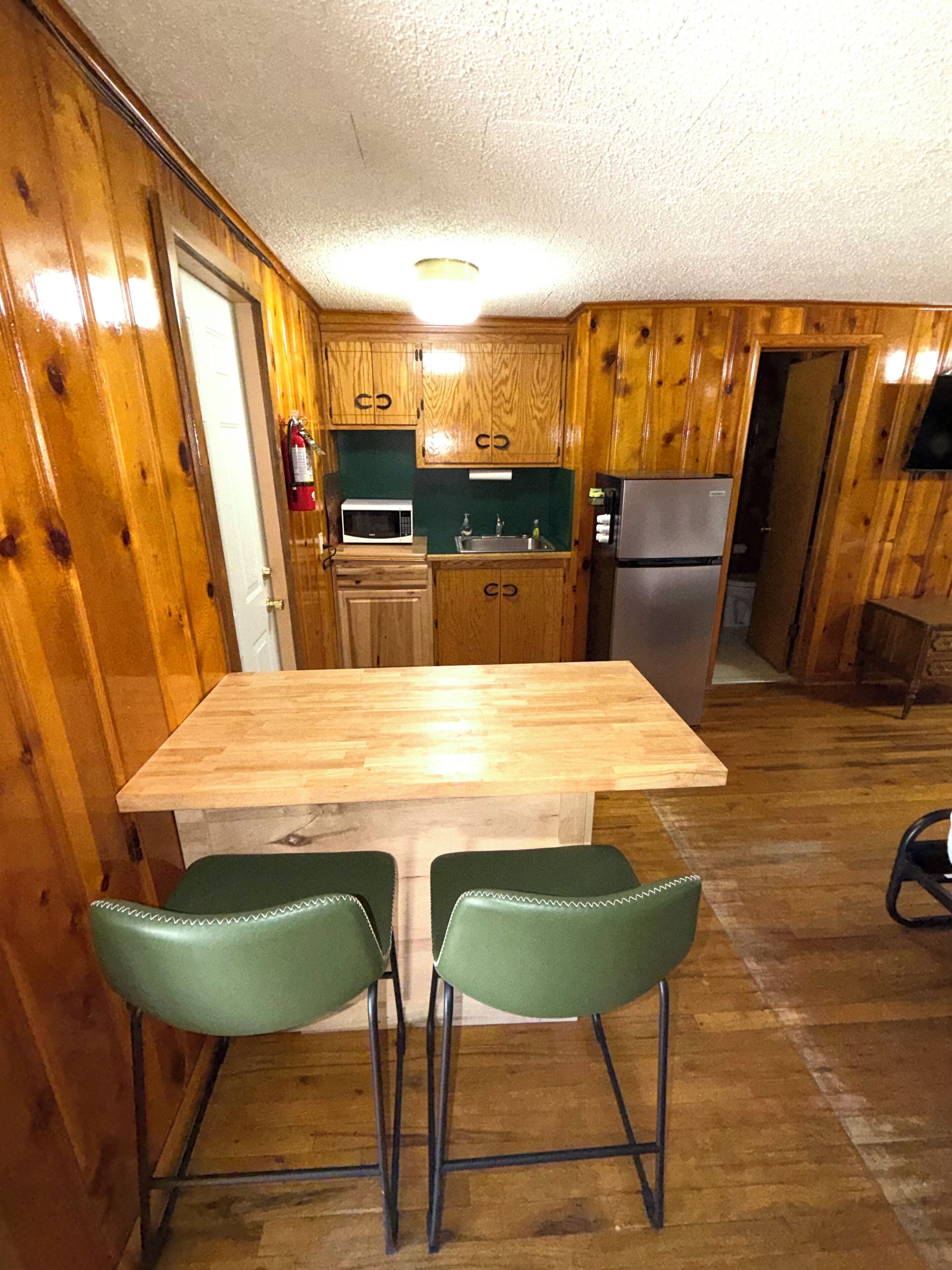 Cocina con paneles de madera, una isla central, dos sillas verdes, un microondas, un fregadero y un refrigerador.