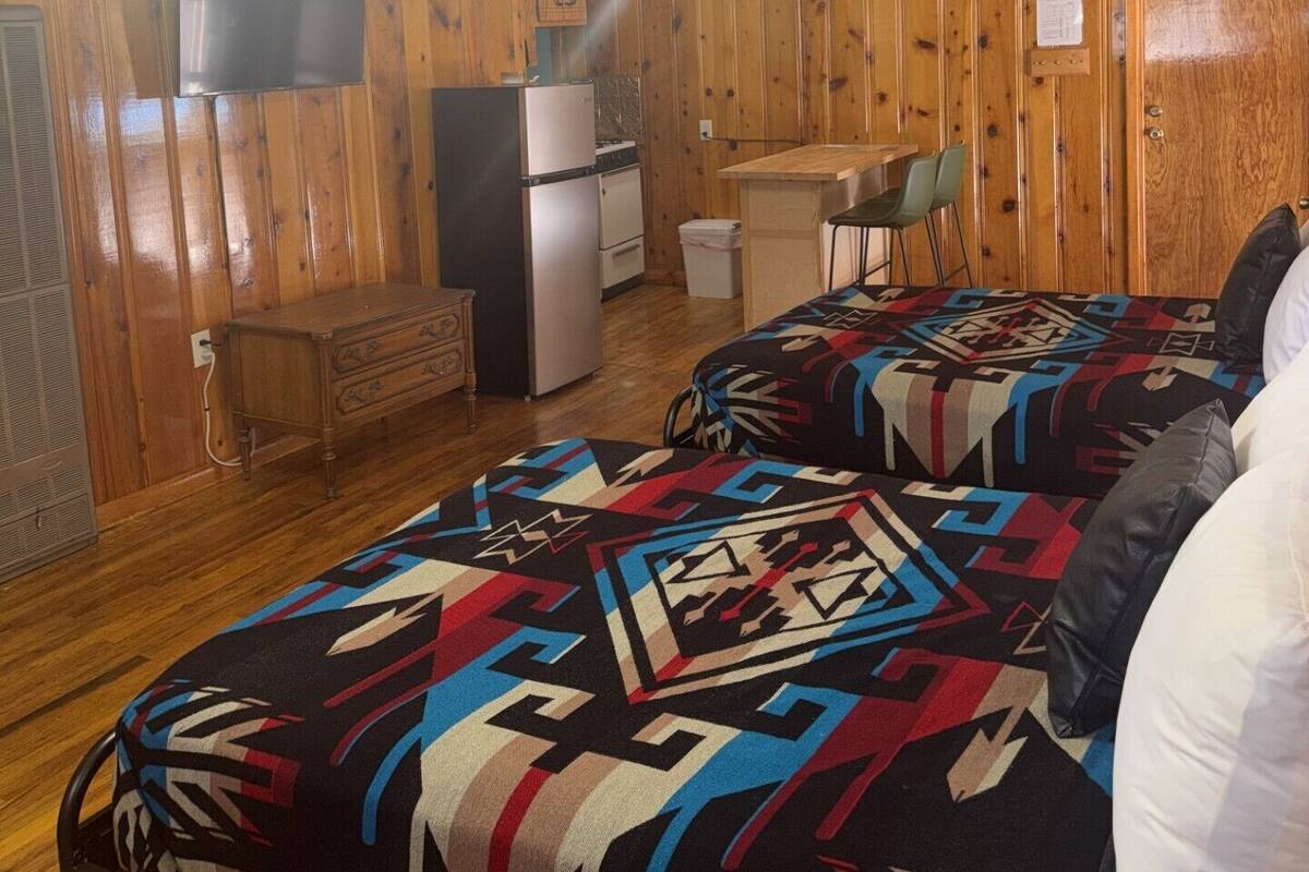 Una habitación con paredes de madera, una cama con una colcha estampada, un escritorio, una silla y un refrigerador.