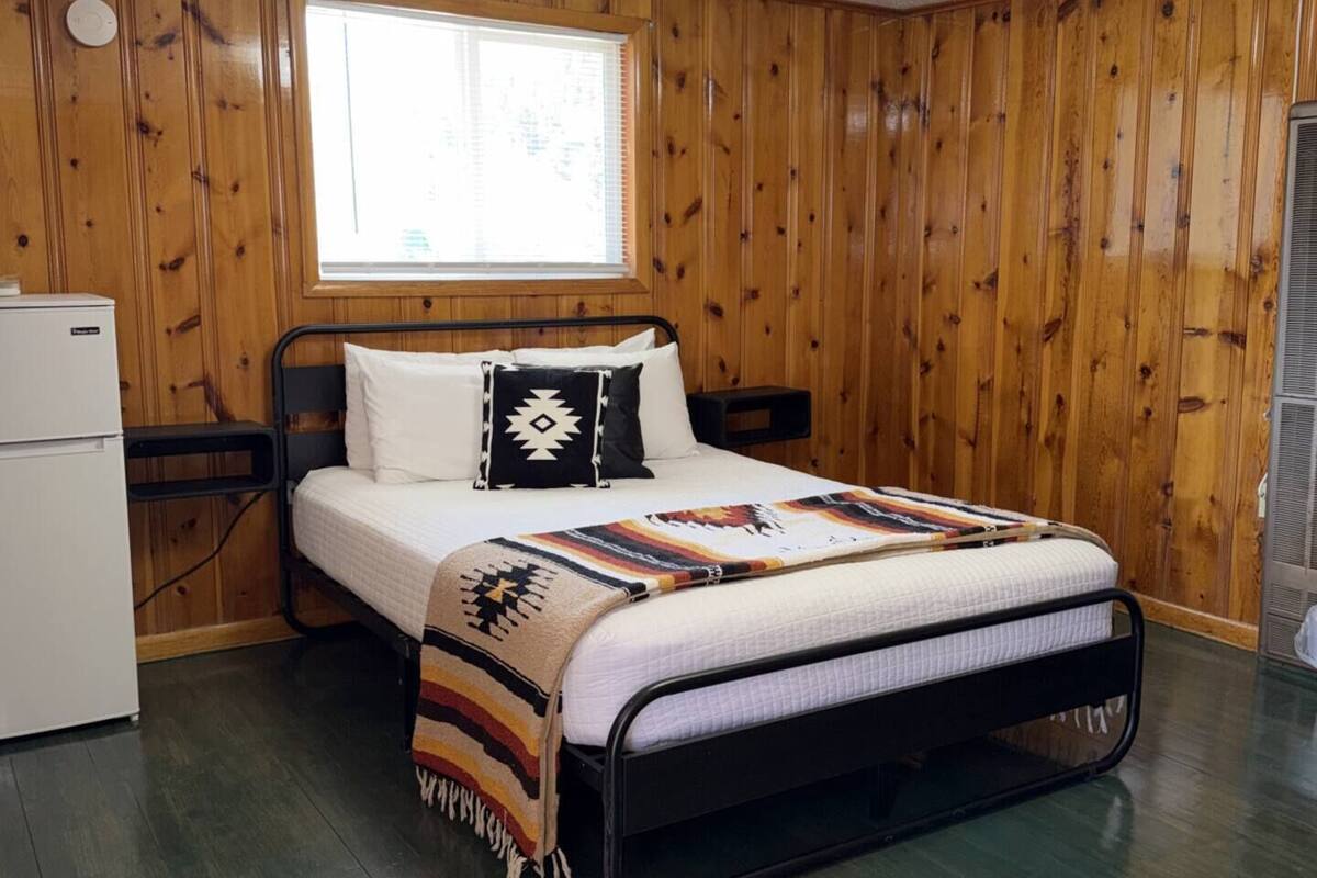 Una habitación con paneles de madera, una cama con colcha estampada, una ventana y un electrodoméstico blanco.