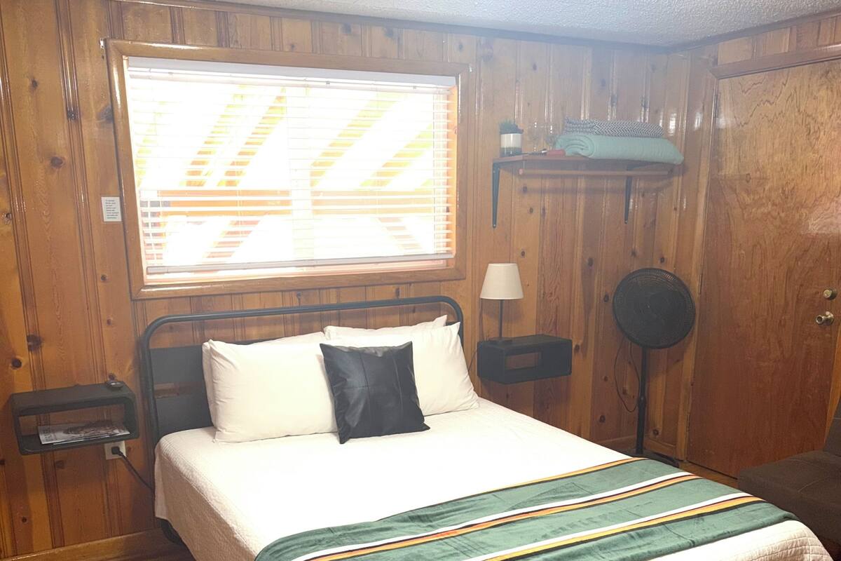 Una habitación con paneles de madera, una cama, una ventana con persianas, una mesita de noche, una lámpara y un aplique de pie.