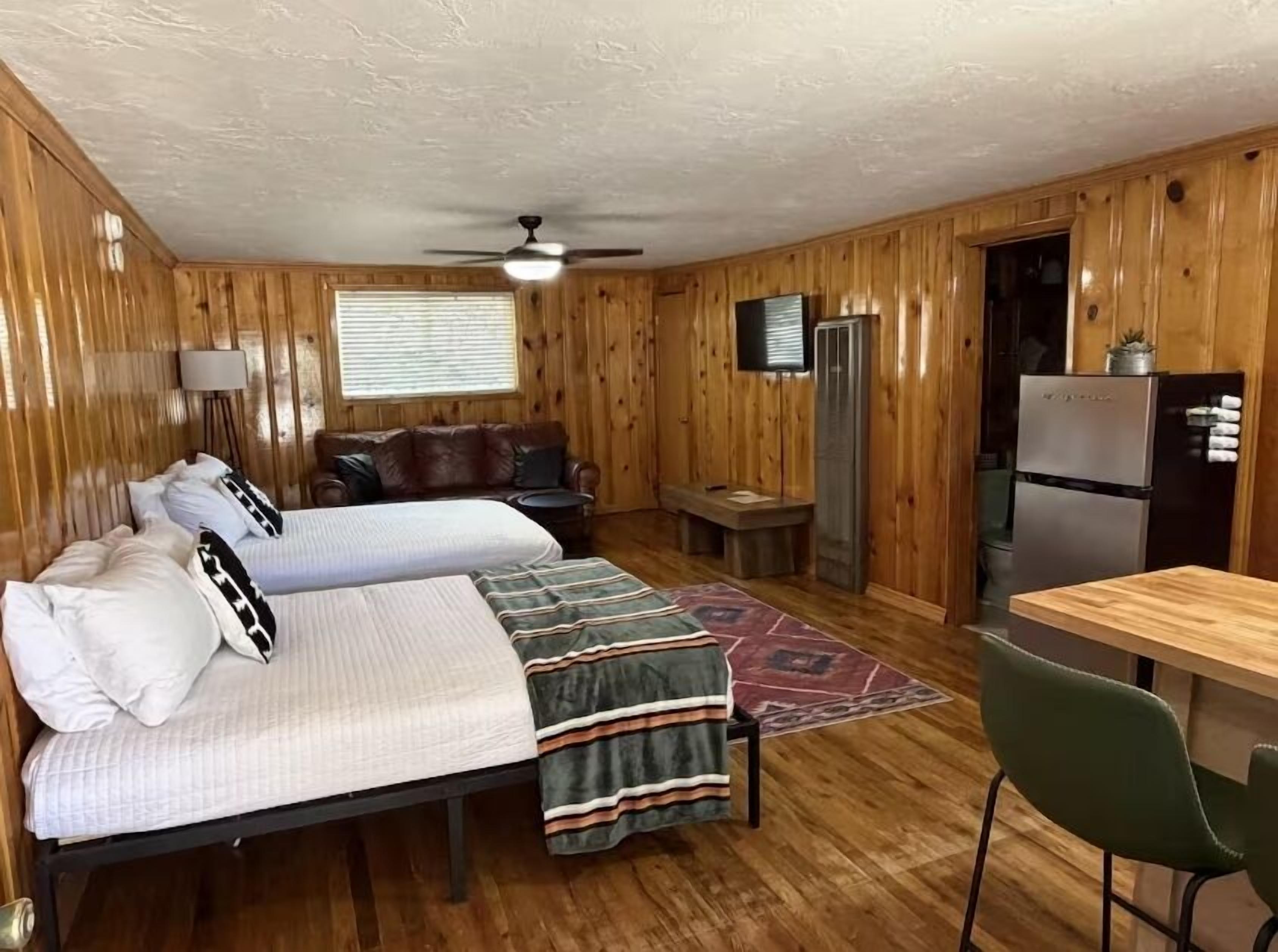 A cabin-style room with two beds, a sofa, a dining table, a refrigerator, and a ceiling fan.