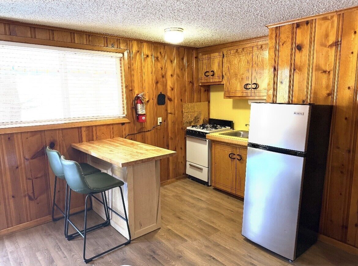A compact kitchen with wooden cabinets, a refrigerator, a stove, and a bar stool.