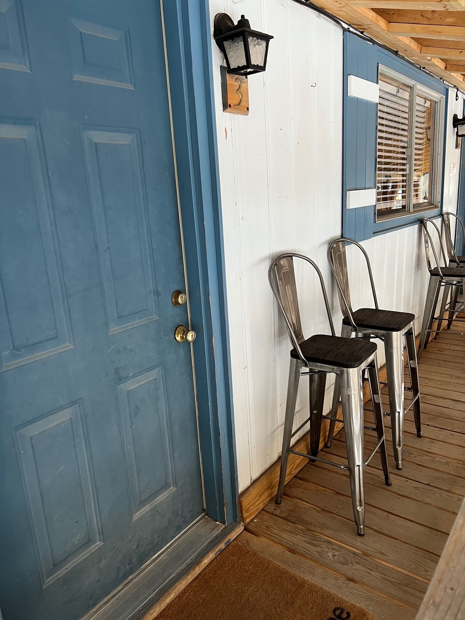 Une porte bleue avec une poignée de porte en laiton, une plaque de numéro en bois et un luminaire noir fixé au mur.