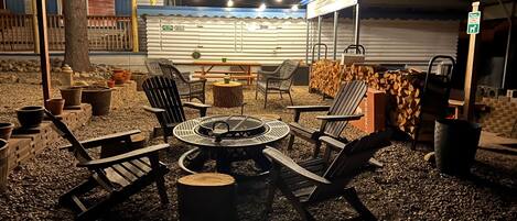 An outdoor seating area with a round table and chairs, surrounded by potted plants and a fire pit.
