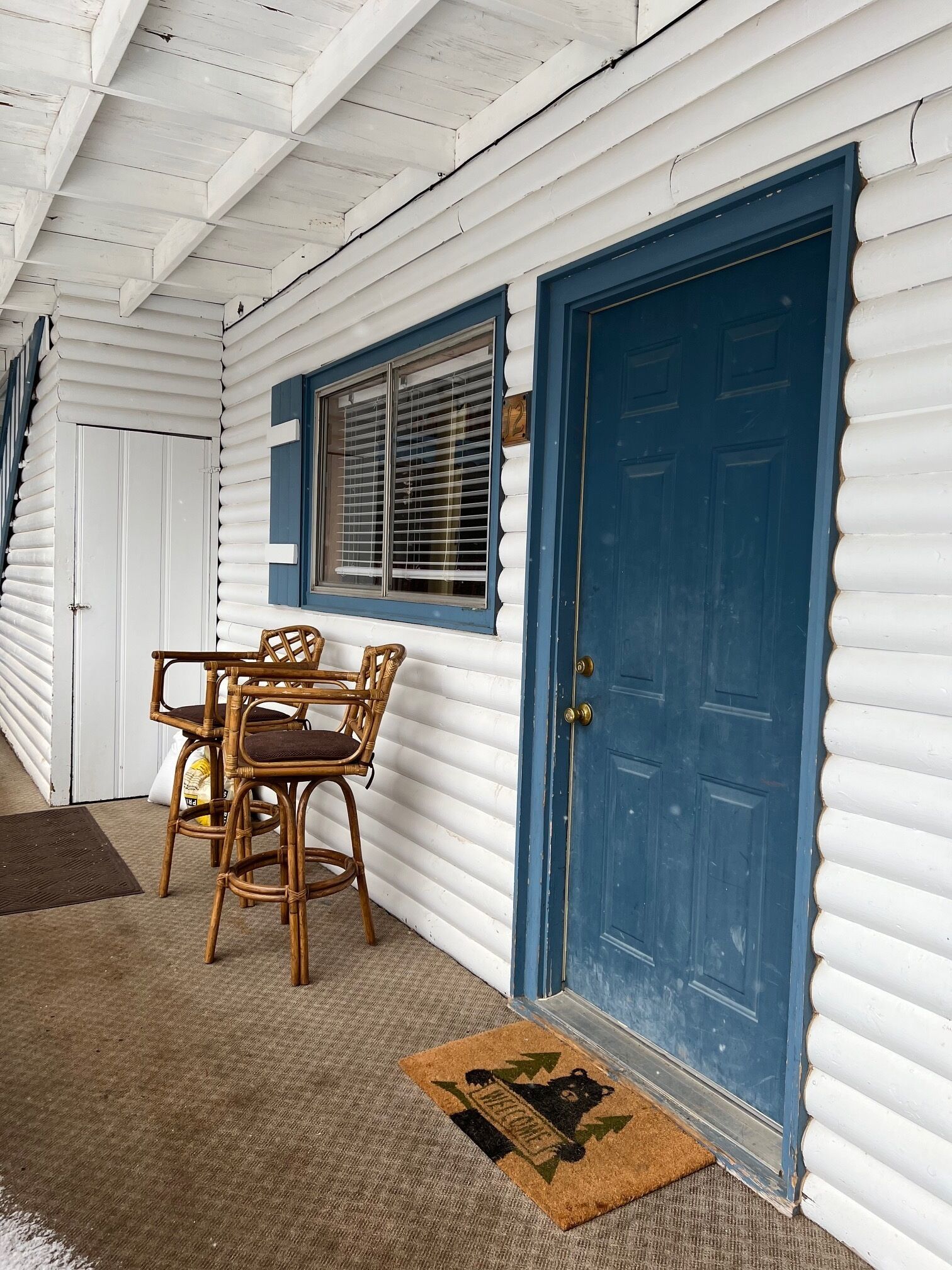 A wooden cabin with a blue door, a window with blinds, and a wicker table with chairs.