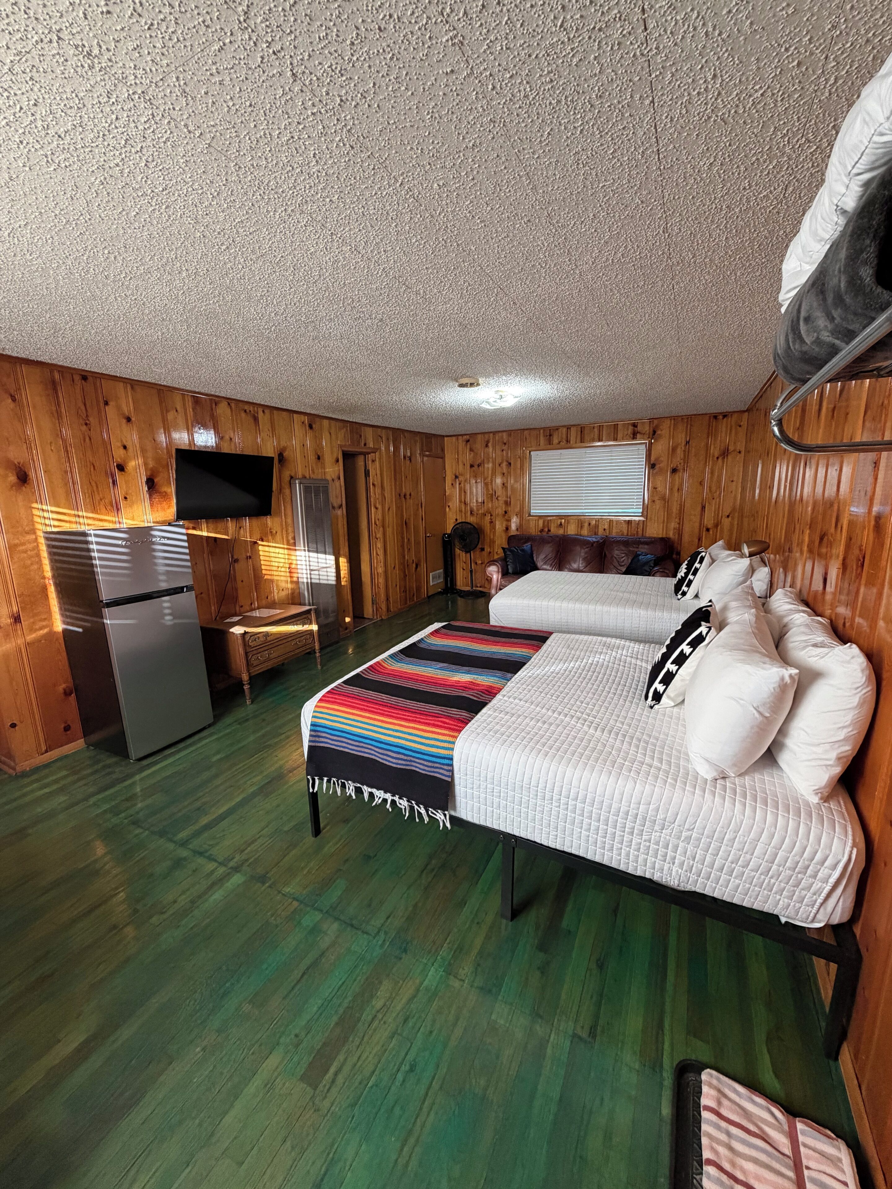 A room with wooden walls, a bed with white bedding, a small kitchenette, and a flat-screen TV mounted on the wall.