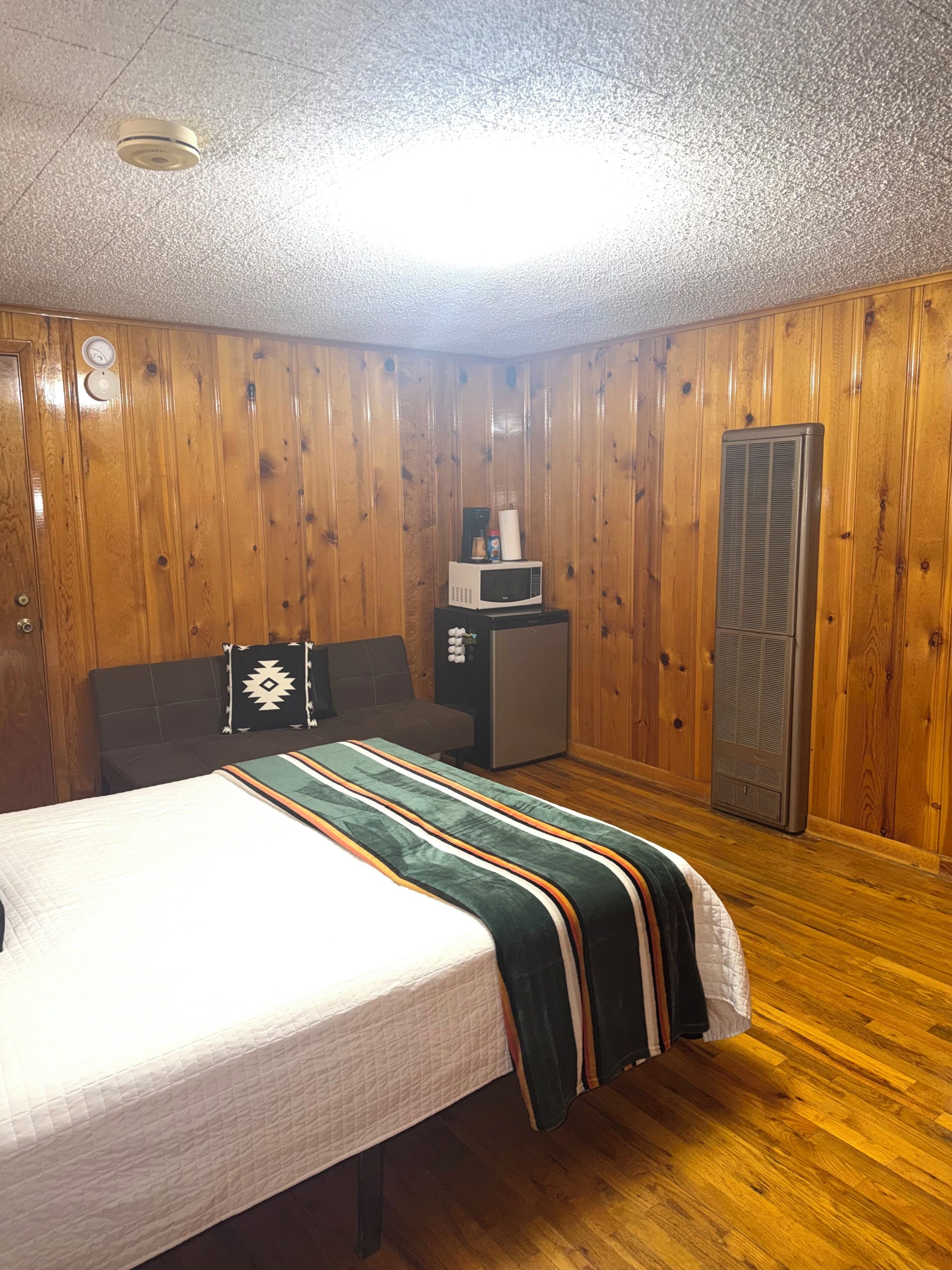 A room with wooden paneling, a bed with a striped blanket, a microwave, a refrigerator, and a heater.