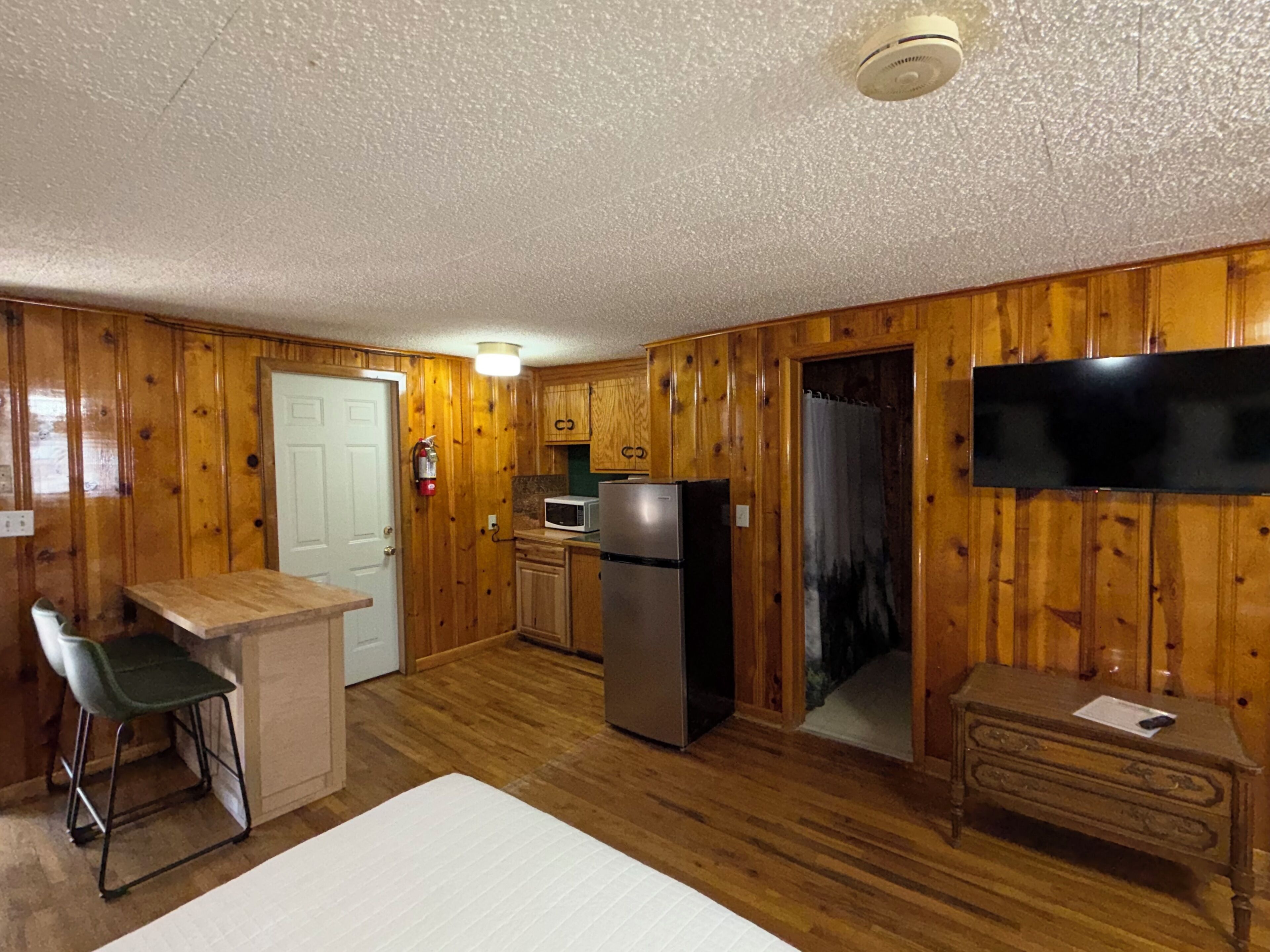 A compact kitchen with wooden walls, a microwave, a refrigerator, a small bar stool, and a flat-screen TV mounted on the wall.