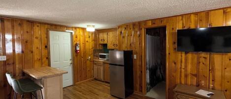 A compact kitchen with wooden walls, a microwave, a refrigerator, a small bar stool, and a flat-screen TV mounted on the wall.