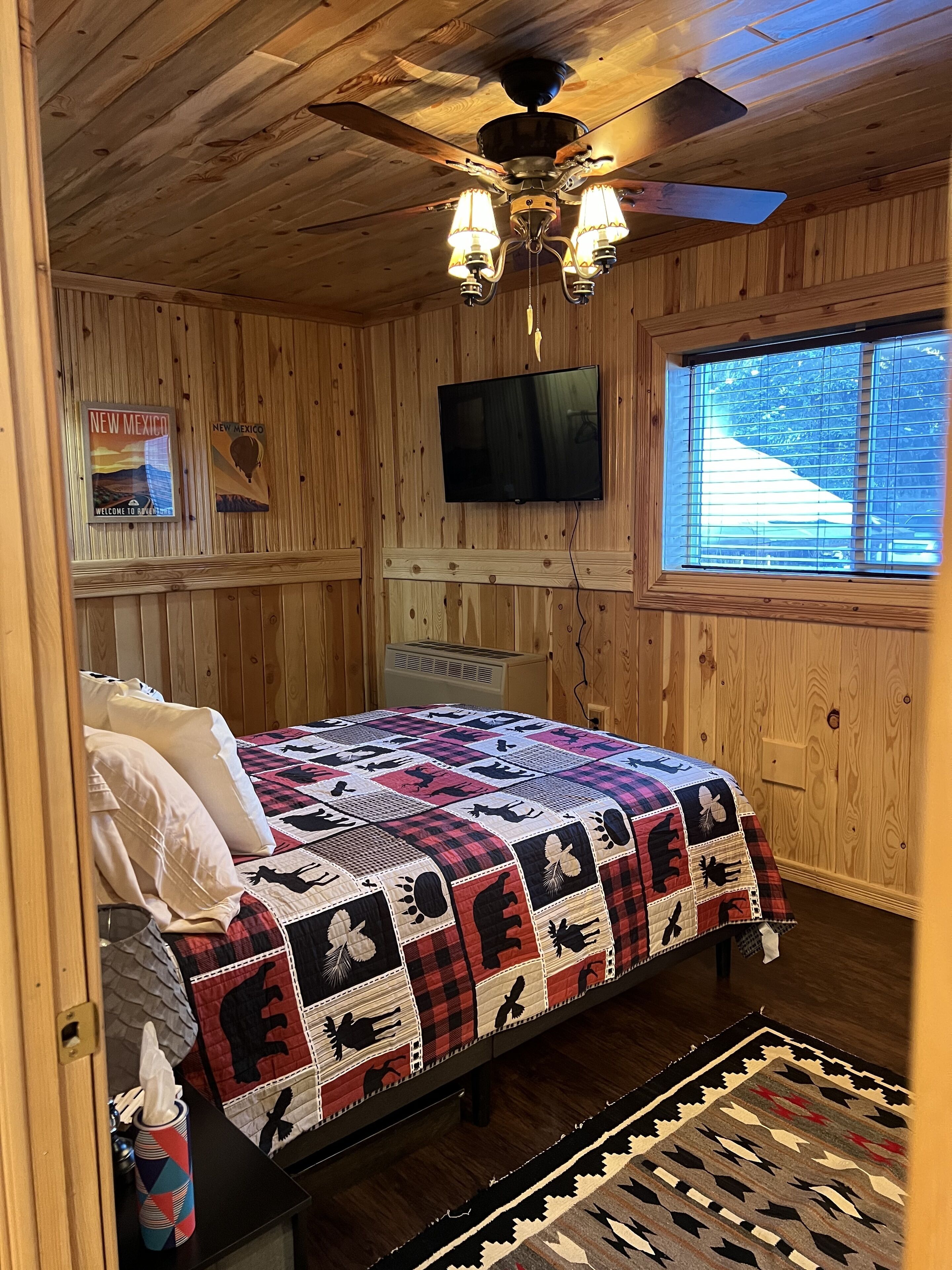 A wooden-paneled room with a bed, a ceiling fan, a mounted TV, and a window with blinds.