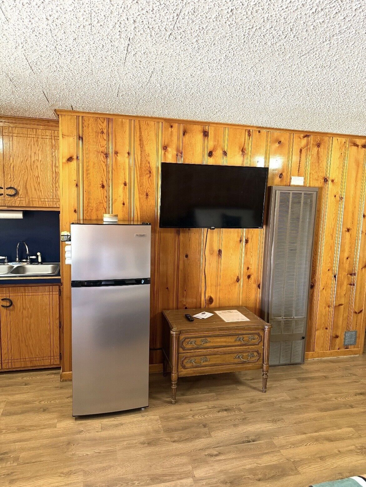 Una cocina con paneles de madera, un refrigerador de acero inoxidable, un televisor de pantalla plana montado en la pared y un aire acondicionado integrado.