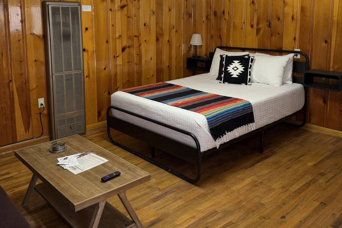 Una habitación con paredes de paneles de madera, una cama con manta a rayas, una mesita de noche con papeles y una lámpara en el techo.