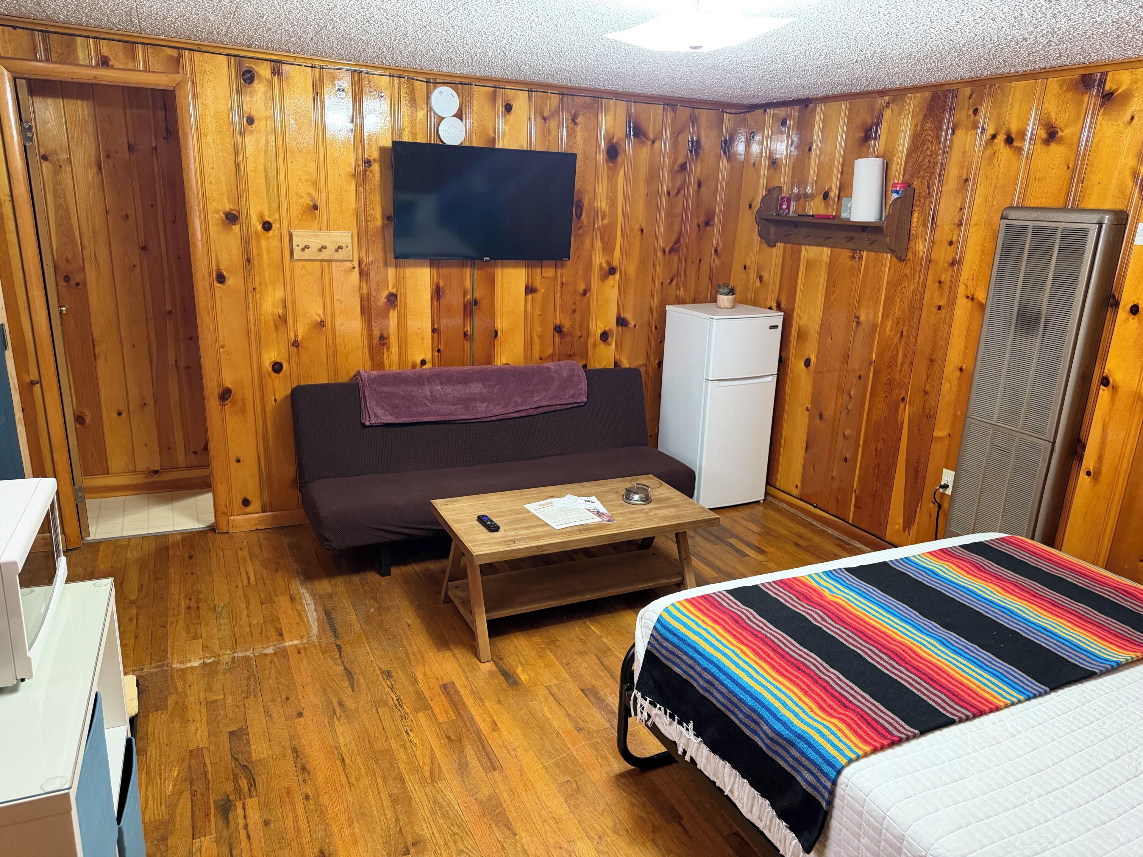 Una habitación con paredes de madera, un sofá, una mesita, una cama con una manta colorida, un televisor, un refrigerador y un calentador.
