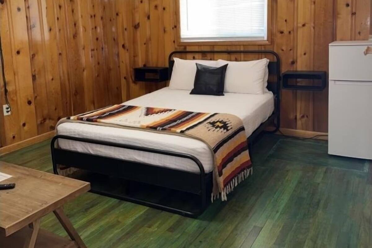 Una habitación con cama, una mesita, una ventana y un refrigerador.