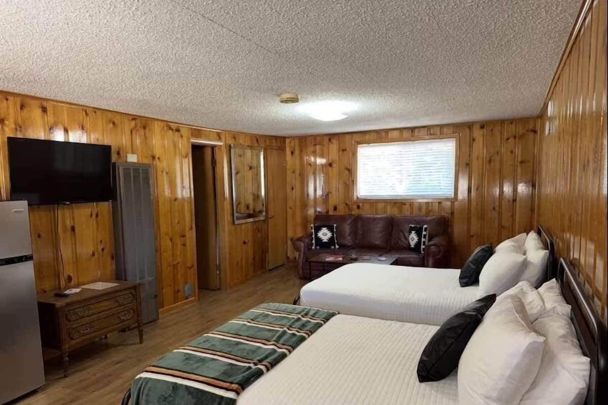 Habitación con paredes de madera, un televisor de pantalla plana, un refrigerador, un aparador, una cama con manta a rayas, un sofá con almohadones y una ventana con persianas.