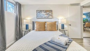 A bedroom with a bed, two bedside lamps, a painting, and a view of the outdoors. - El Sereno Bungalow! Pet-Friendly Ivywild Flat (Colorado Springs)