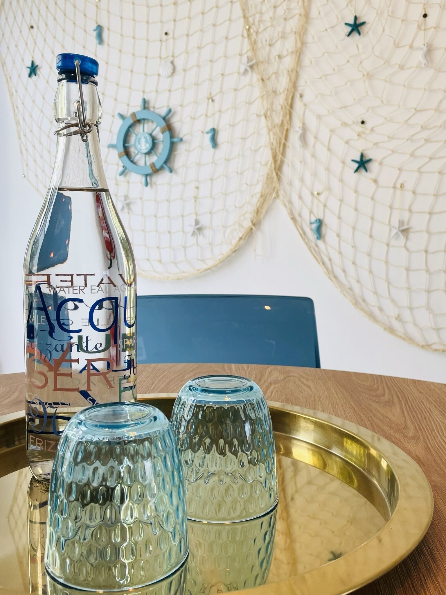 Une bouteille en verre transparent avec un bouchon bleu et une étiquette, posée sur un plateau avec deux verres.