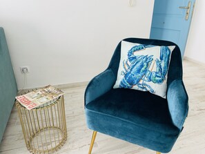 Una poltrona blu scuro con un cuscino a forma di aragosta blu e bianca, un tavolino da caffè in filo d'oro con riviste, e una porta blu sullo sfondo.