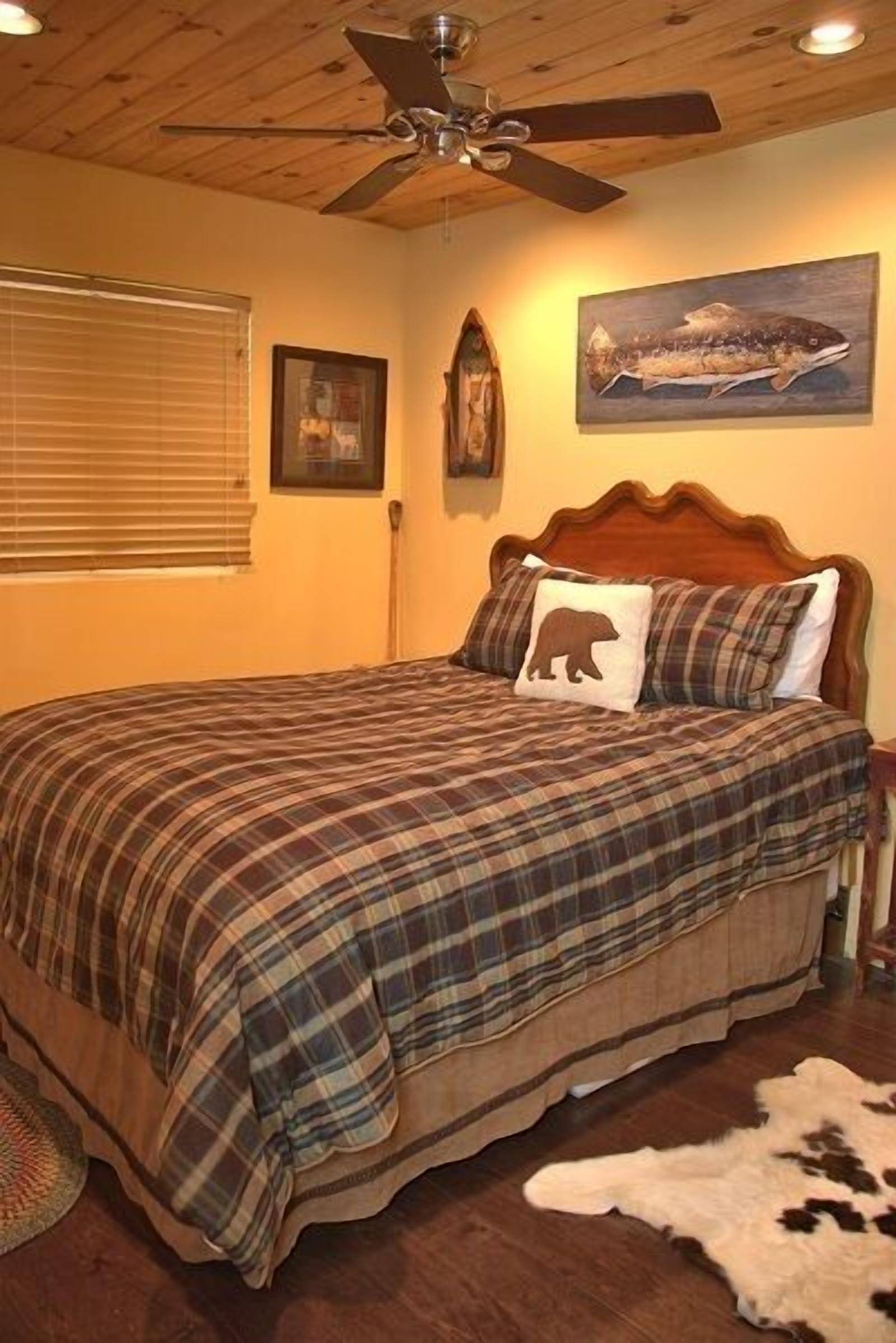 A bedroom with a plaid bedspread, a wooden headboard, a ceiling fan, a framed picture of a fish, and a fur rug.