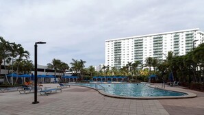 Zona de piscina con sillas de descanso y sombrillas, rodeada de palmeras y un edificio de hotel alto al fondo.
