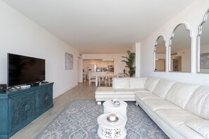 A modern living room with a large flat-screen TV, a white leather sofa, and a dining area in the background.