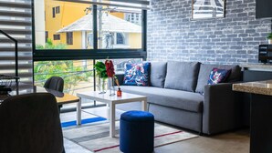 A modern living room with a grey sofa, a blue ottoman, and a white coffee table. A large window provides a view of a yellow building outside. - New Loft NYC style, King Bed, High Speed Wifi, (Santo Domingo)