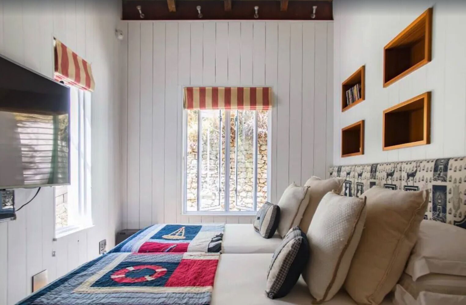 Une chambre avec une grande fenêtre, un lit recouvert d’une courtepointe, un téléviseur et des étagères fixées au mur.