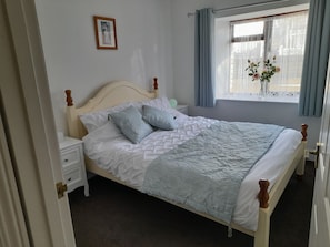 A neatly made bed with a white headboard and wooden footboard, a bedside table with a lamp, and a window with curtains.