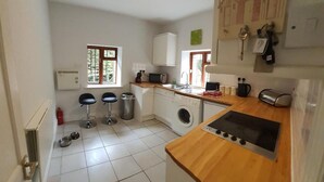 A compact kitchen with a washing machine, a microwave, and a stove. There are two stools and a window with a view of greenery. - Cosy Apartment Close to Tunnels Beaches (Ilfracombe)