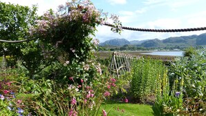 A garden with various plants and flowers, a rope bridge, and a view of mountains and a lake in the background. - Lodge 3 - Pet Friendly - Aultbea Lodges - Sea Views of Loch Ewe (Achnasheen)