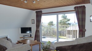 A living room with a sofa, a coffee table, a TV, and a balcony with a railing. - Lodge 3 - Pet Friendly - Aultbea Lodges - Sea Views of Loch Ewe (Achnasheen)