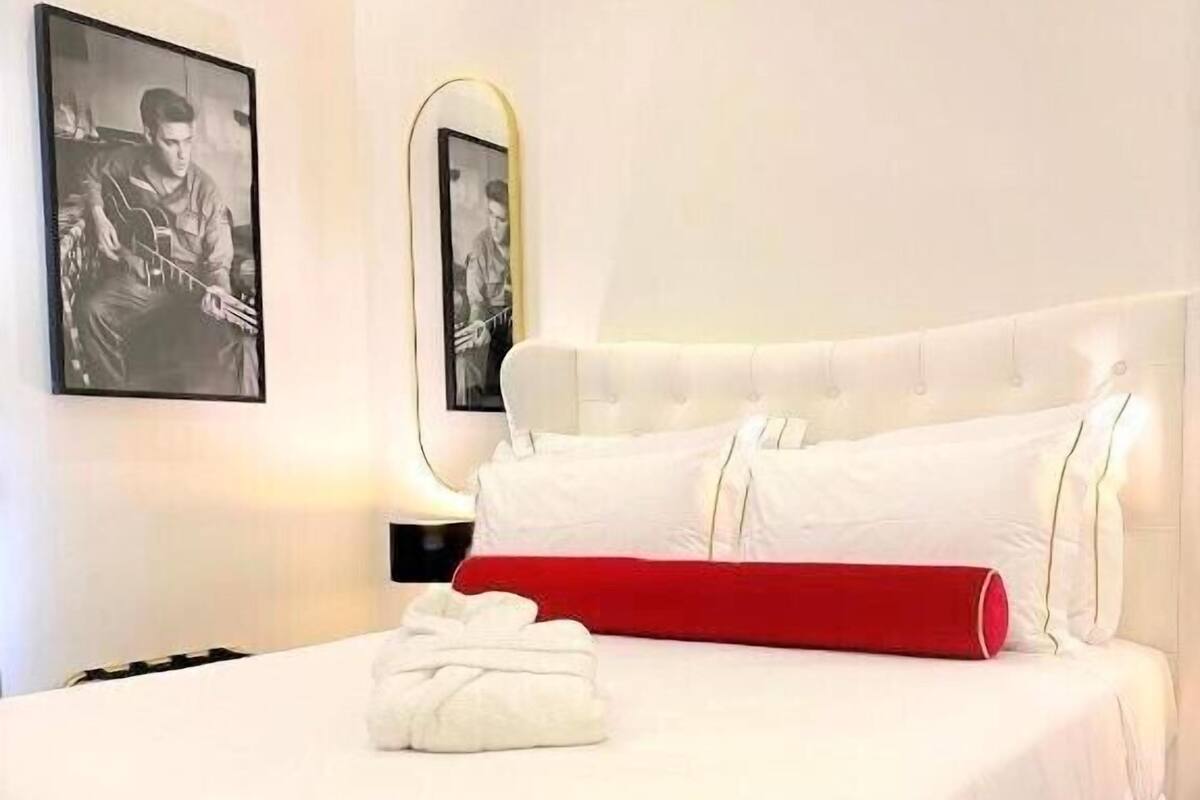 Ein ordentlich bezogenes Bett mit weißer und roter Bettwäsche, ein weißer Bademantel auf dem Bett sowie zwei gerahmte Bilder an der Wand.