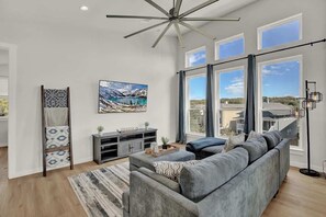 A spacious living room with a large gray sectional sofa, a modern ceiling fan, and a large window offering a view of the outdoors.