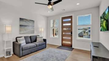 Una sala de estar moderna con un sofá gris, un ventilador de techo y ventanas grandes con persianas.