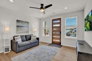A modern living room with a gray sofa, a ceiling fan, and large windows with blinds.