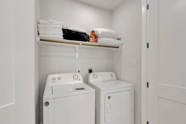 A laundry room with two white washing machines and a shelf above holding folded towels and a detergent bottle.