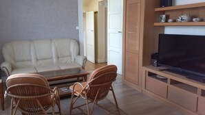 A living room with a sofa, two wicker chairs, a coffee table, a TV on a wooden stand, and a door in the background.