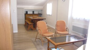 A dining area with a glass-top table, two wicker chairs, a wooden bench, and a kitchen in the background.