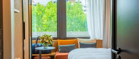 A compact hotel room with a bed, a small seating area with a table and chairs, and a large window offering a view of trees and buildings.