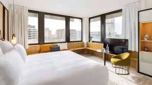 Habitación de hotel con una cama grande, zona de estar con televisor y vistas a la ciudad.