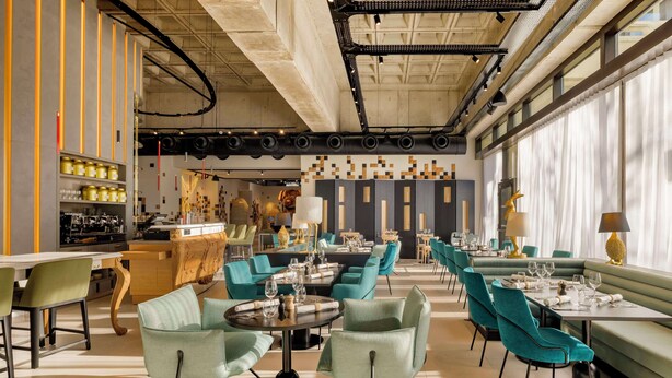 Interior moderno de restaurante con sillas color turquesa, mesas redondas y una zona de bar con estantes llenos de botellas.