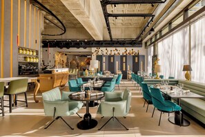 Interior moderno de restaurante con sillas color turquesa, mesas redondas y una zona de bar con estantes llenos de botellas.
