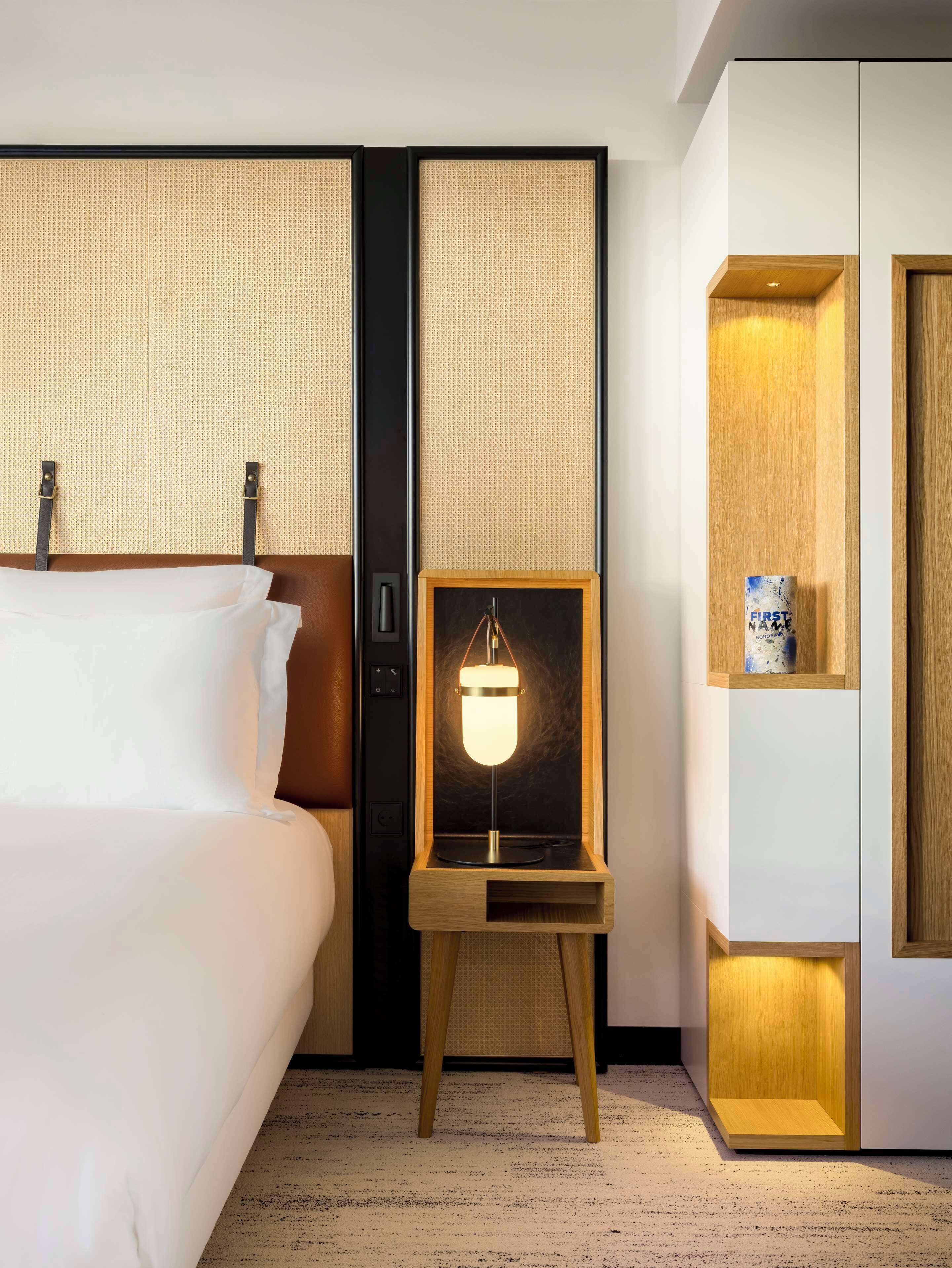 A hotel room with a bed, a nightstand with a lamp, and a wooden cabinet with a small shelf.