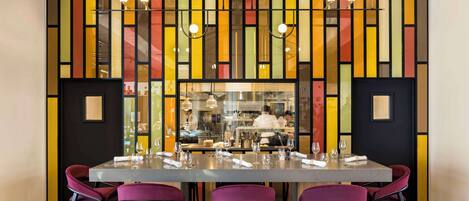 Un espace repas avec une longue table, des chaises violettes et un mur rayé aux couleurs vives.