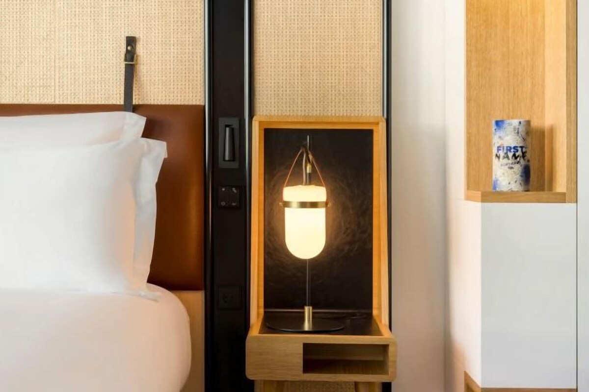Una habitación de hotel con una cama, una mesita de noche con lámpara y un estante de madera con un objeto decorativo.